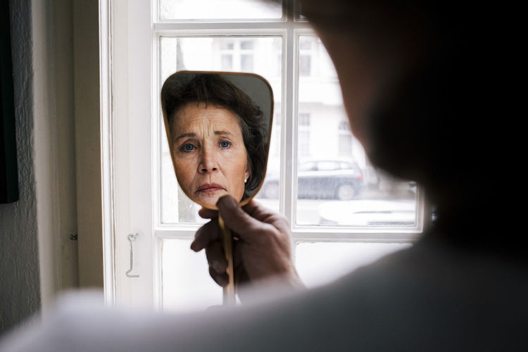 Reflection of worried retired senior woman with wrinkles on hand mirror at home
