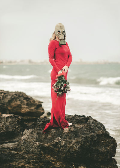 Woman wearing gas mask holding bouquet standing on rock at beach