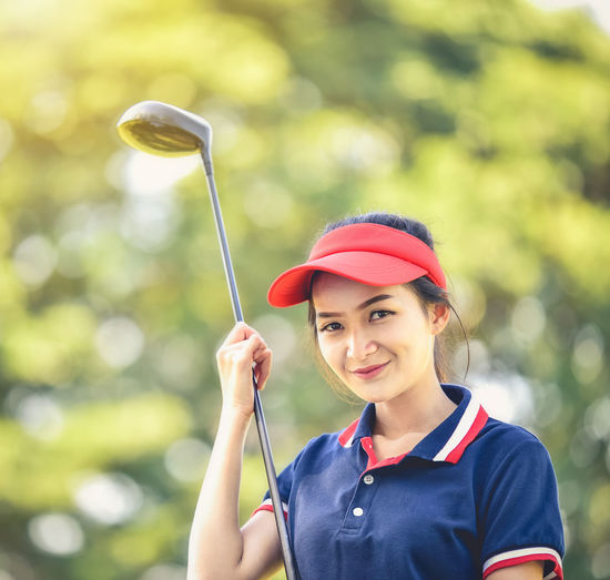 Portrait of smiling young woman holding golf club against tree