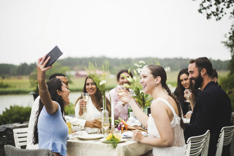 Cheerful young woman taking selfie with friends during dinner party in backyard