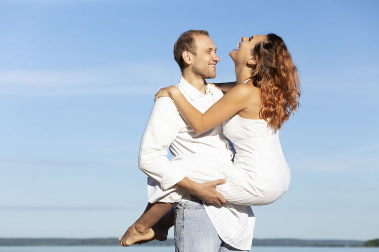Happy smiling caucasian man carries laughing latin woman on hands, blue sky,water on background