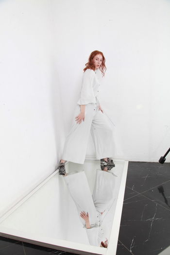 Full length fashion catalog style portrait of woman in jumpsuit standing on mirror over white wall