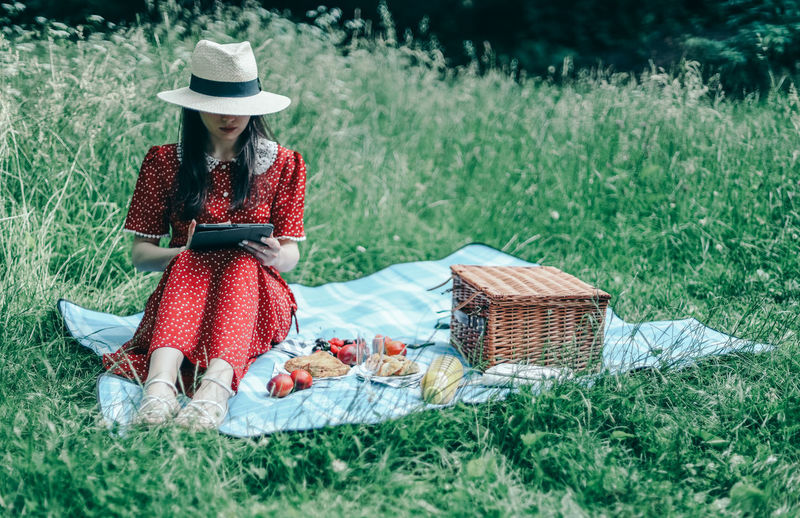 A beautiful young caucasian girl in a red dress sits on a blue bedspread and reads an e-book