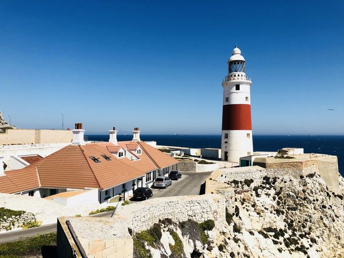 Lighthouse by sea against buildings against clear blue sky