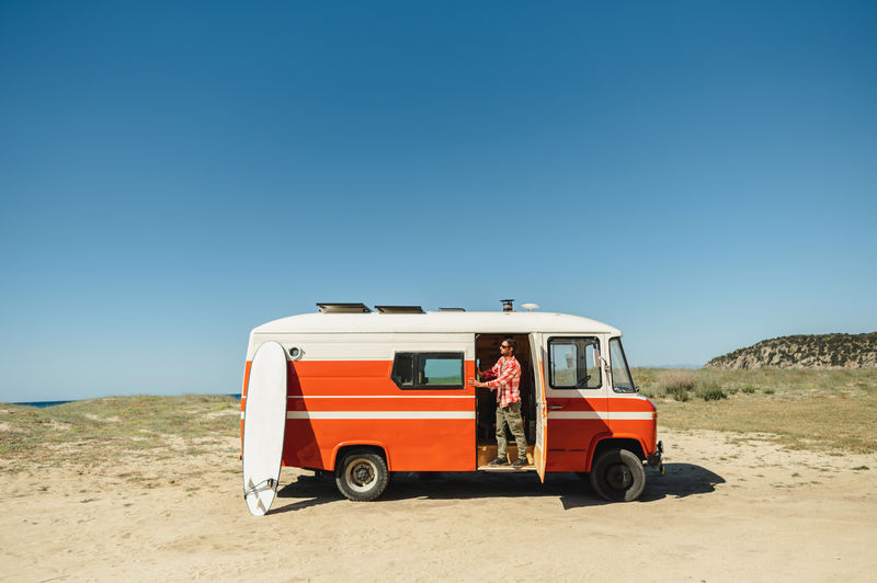 Full body of young male tourist in casual clothes and sunglasses standing in vintage camper van parked on sandy beach before surfing