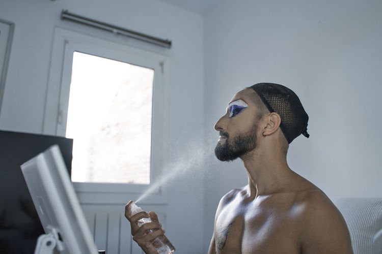 Shirtless man wearing hair net setting make-up with spray