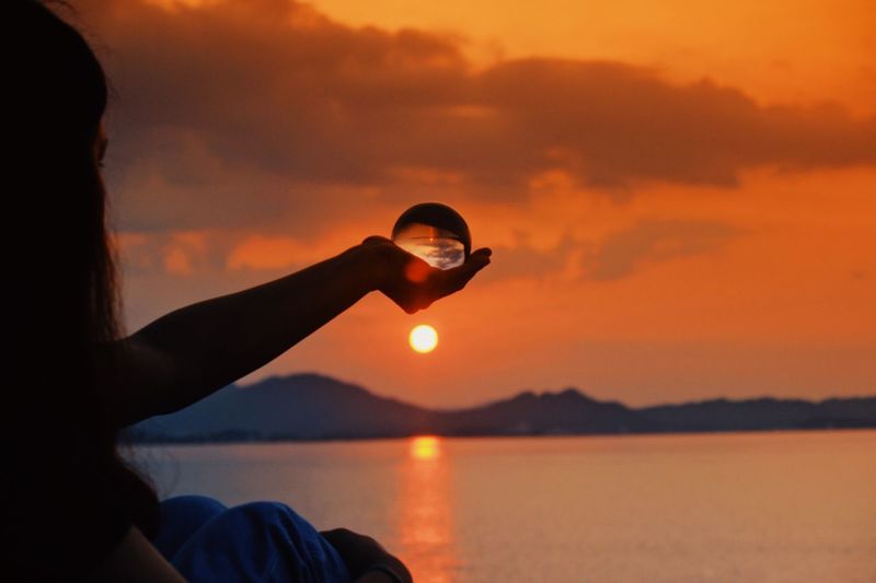 Woman holding crystal ball against sea during sunset
