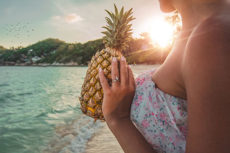 Midsection of woman holding pineapple at beach against sky