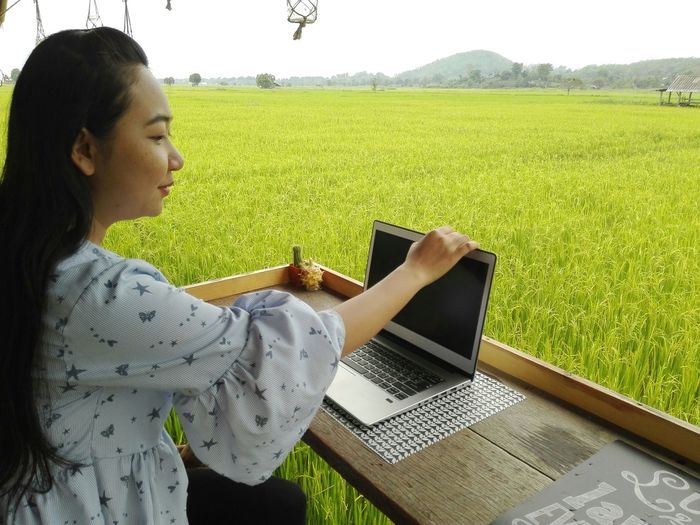 Woman using laptop while sitting at table against farm