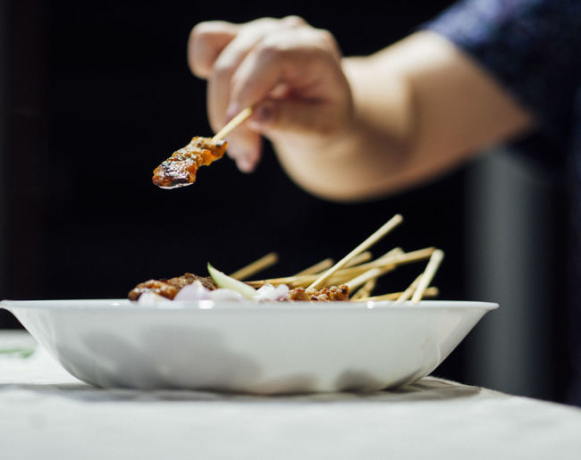 Hand holding stick of satay above dish of satay