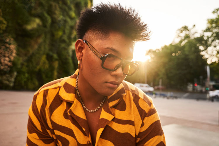 Confident asian male wearing cool shirt with sunglasses and accessories looking at camera while standing in sundown light on street