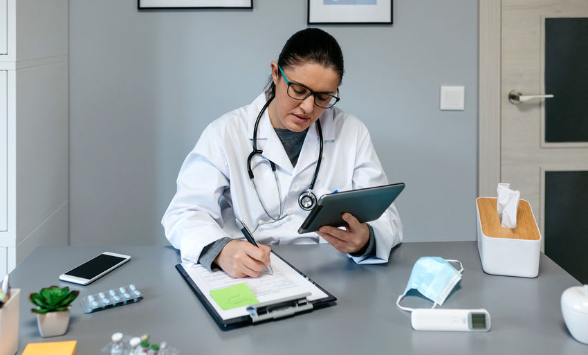 Doctor using digital tablet while writing on document at desk in office