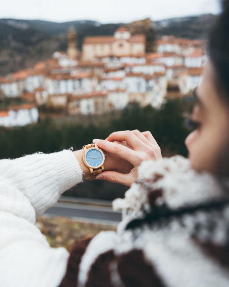 Back view of crop female traveler checking time on trendy wristwatch while standing on hill against old town in cloudy autumn day