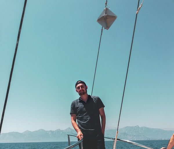Young man standing by railing of boat in sea against clear sky