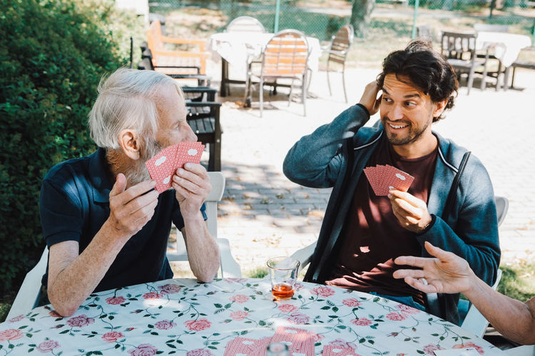 Smiling male caretaker playing cards with senior man and woman at table in back yard