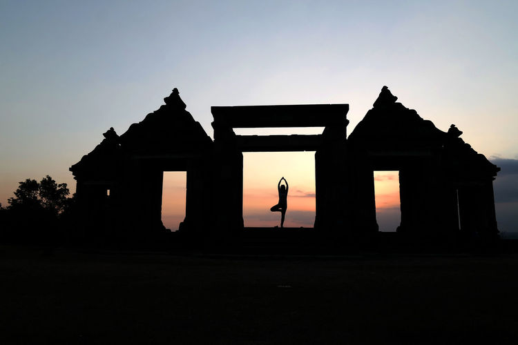Silhouette person performing yoga at old built structure against sky during sunset