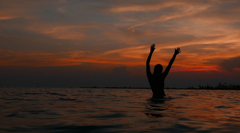 Silhouette woman with arms raised swimming in sea against sky during sunset