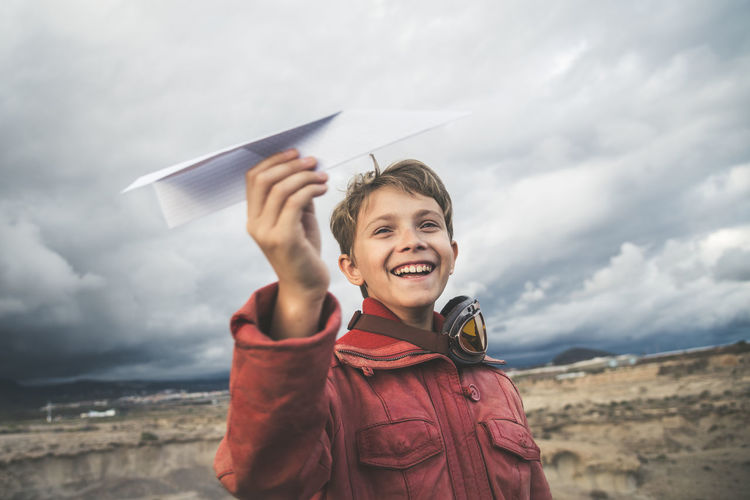 Smiling boy holding paper airplane while standing against cloudy sky