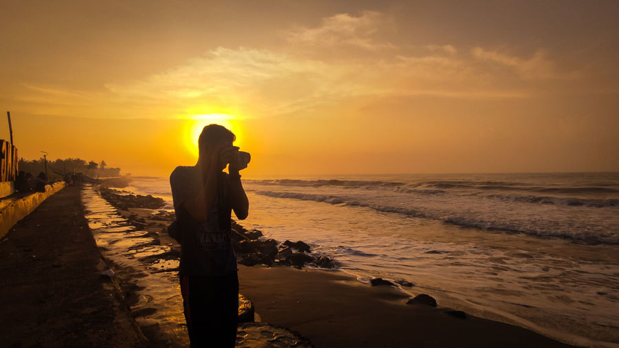 Man photographing while standing on beach against sky during sunset