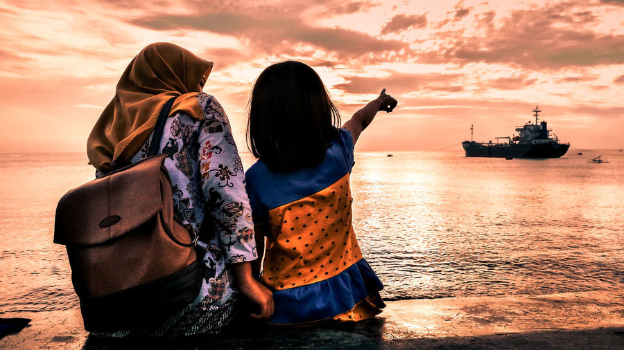 Girl pointing while sitting by mother at beach against sky during sunset