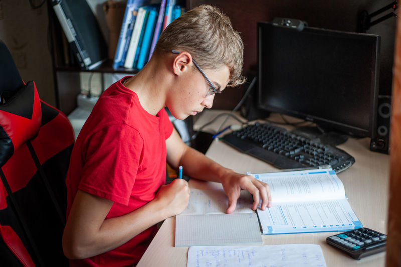 A teenage boy in a red t-shirt sits at a table at home and does his homework. study without gadgets.