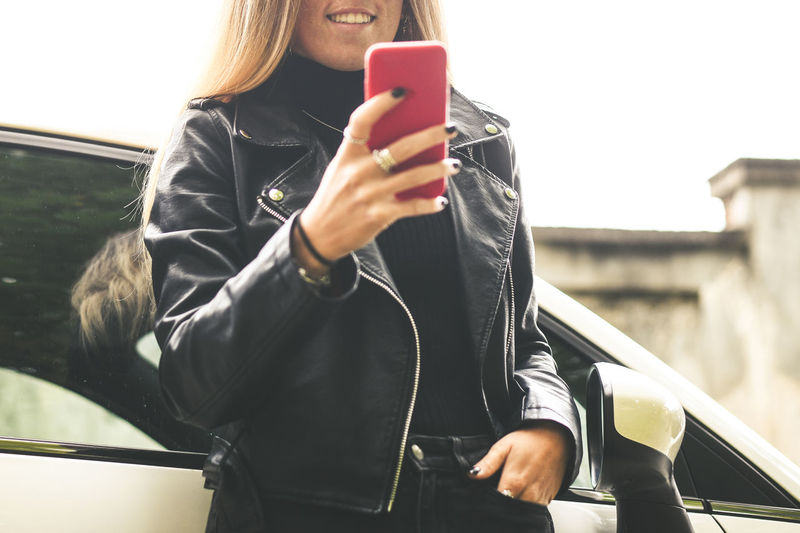 Trendy blonde girl using smartphone leaning against her car. teen use mobile phone chat with friends