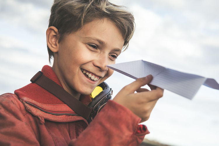 Smiling boy playing with paper airplane against cloudy sky