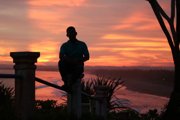 Man sitting on railing against sky during sunset