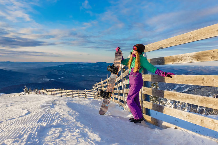 Full length if woman standing by wooden fence with snowboard against sky