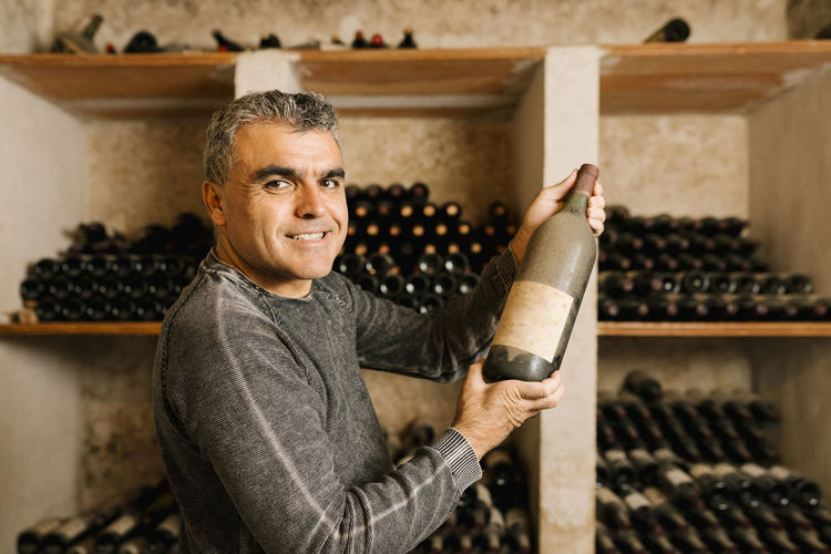 Smiling male owner holding old wine bottle while standing by rack at cellar