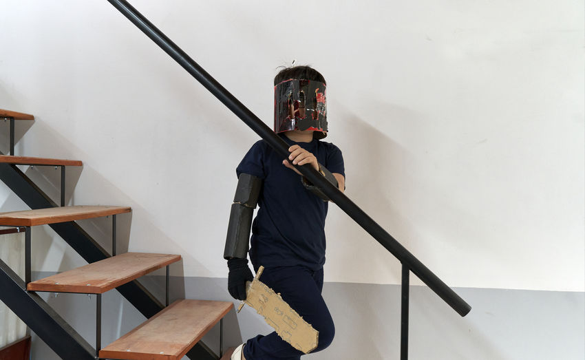 Cute boy wearing cardboard mask and holding sword standing on staircase at home