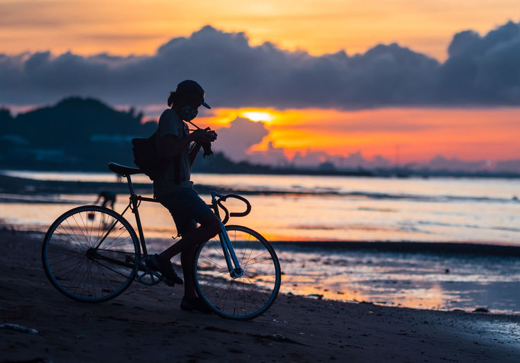 Man on bicycle at beach against sky during sunset