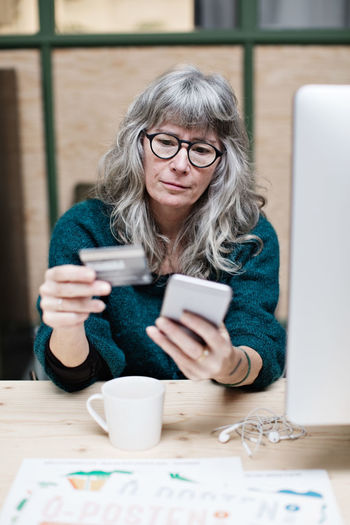 Mature businesswoman shopping online through mobile phone while having coffee at office