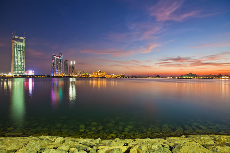 Illuminated buildings by river in city against sky during sunset