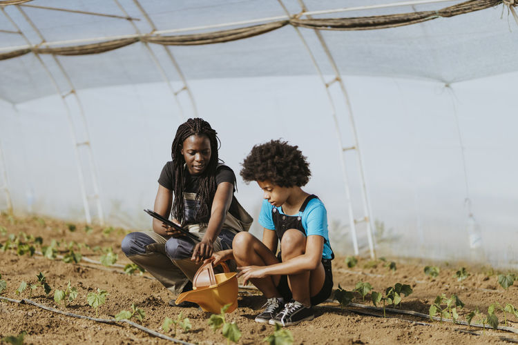 Female farmer teaching gardening to afro girl while crouching at greenhouse
