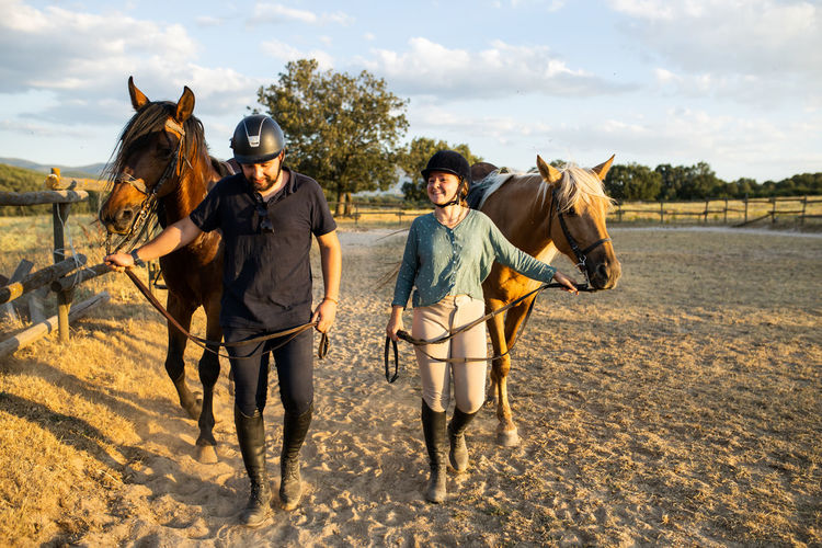 Smiling female and male trainer in protective helmets strolling with stallions on sandy terrain under cloudy sky