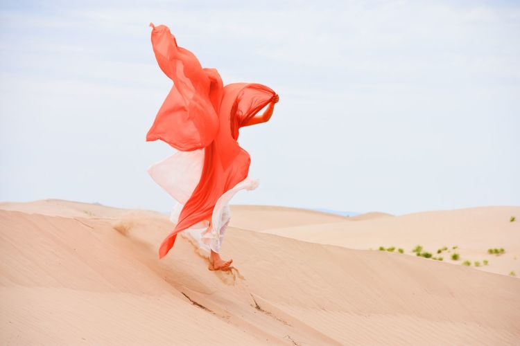 Person on sand dune in desert against sky wearing large pink coral scarf 