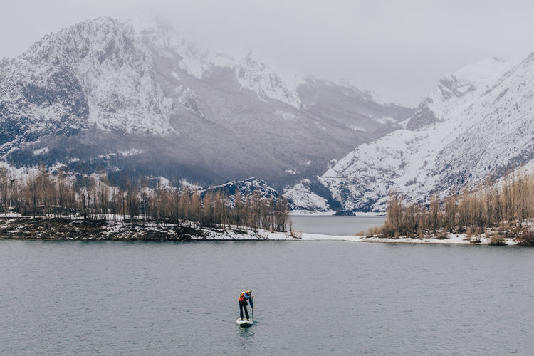 Person floating on paddle board between water surface and picturesque view of hills in snow