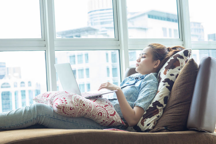Young woman using mobile phone while sitting on window