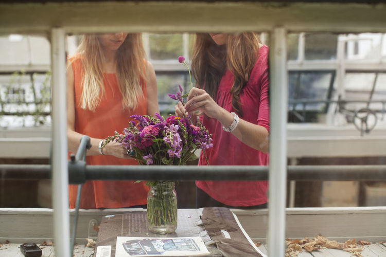 Midsection of women putting flowers in jar seen through glass