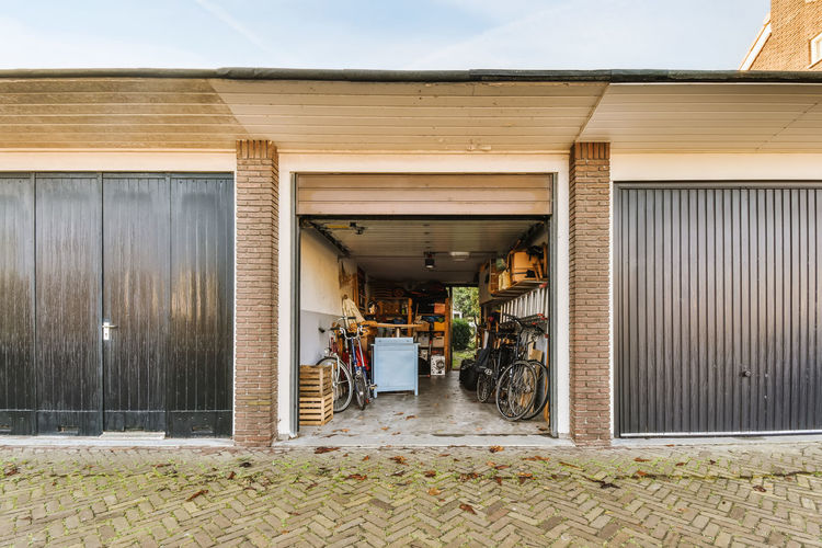 Opened metal garage with parked bicycles and wooden boxes with various equipment on shelves located in countryside on summer day