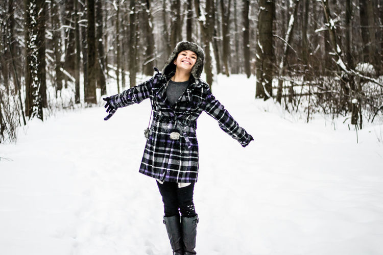 Smiling young woman standing with arms outstretched against trees in forest during winter