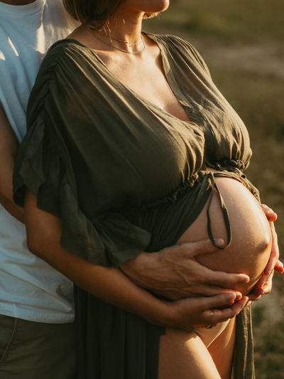 Cropped unrecognizable male hugging pregnant female from behind while standing in countryside meadow
