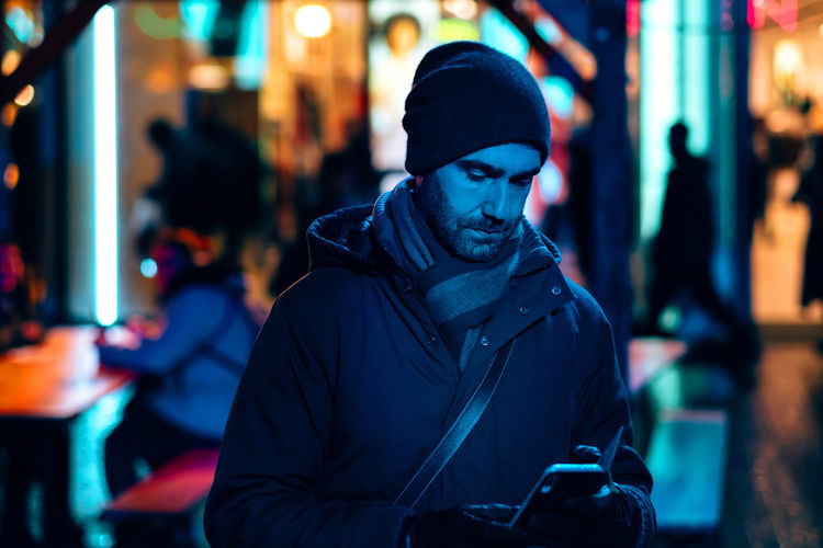 Portrait of young man with phone standing against illuminated city at night