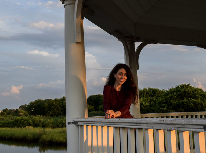Portrait of smiling young woman standing by railing against sky