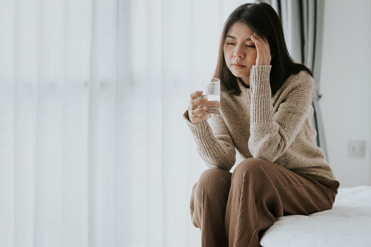 Woman with headache holding glass while sitting on bed at home