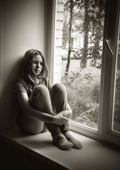 Depressed girl hugging knees on window sill at home