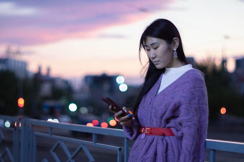 Asian woman using mobile phone in urban scene during sunset on blurred background