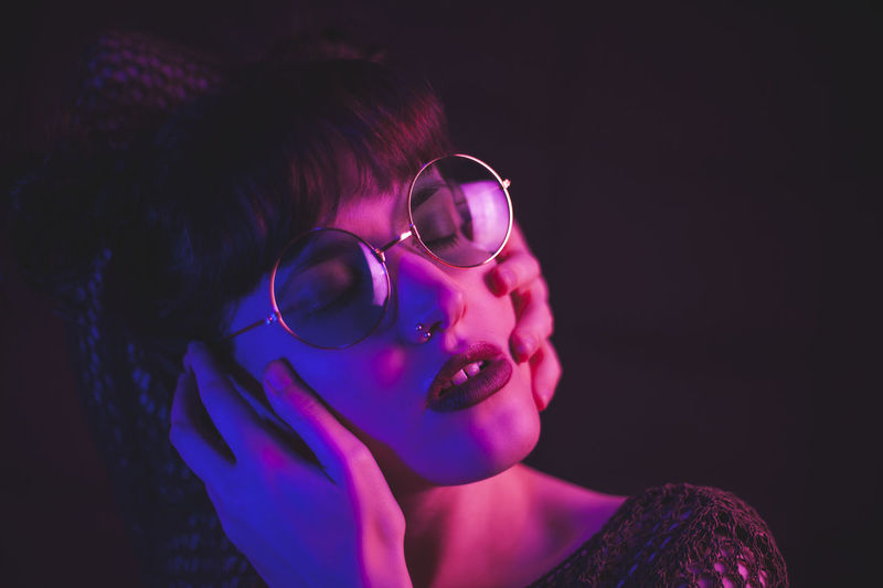Tranquil female in stylish glasses touching face while standing with closed eyes on dark background in pink and blue neon illumination