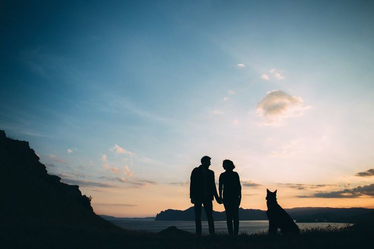 Silhouette couple with dog standing at beach against sky during sunset